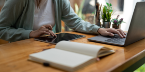 Woman prepares for a meeting by using a laptop and tablet at a wooden desk with a notebook and potted plants.