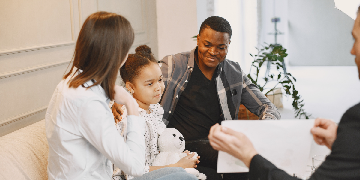 Smiling parents sit with their daughter and a mediator during a calm family meeting, with the child holding a white stuffed animal.